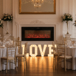 Kominki elektryczne w ślubnych aranżacjach. Trend sezonu czy przemyślana inwestycja w niezapomnianą atmosferę? Kominkek Z Obudową Na Ślub. Kominek elektryczny na sali weselnej. Biała sala z kominkiem led na centralnej ścianie. Pod kominkiem podpis neonowy LOVE. Aranżacja sklep online kominki elektryczne.pl